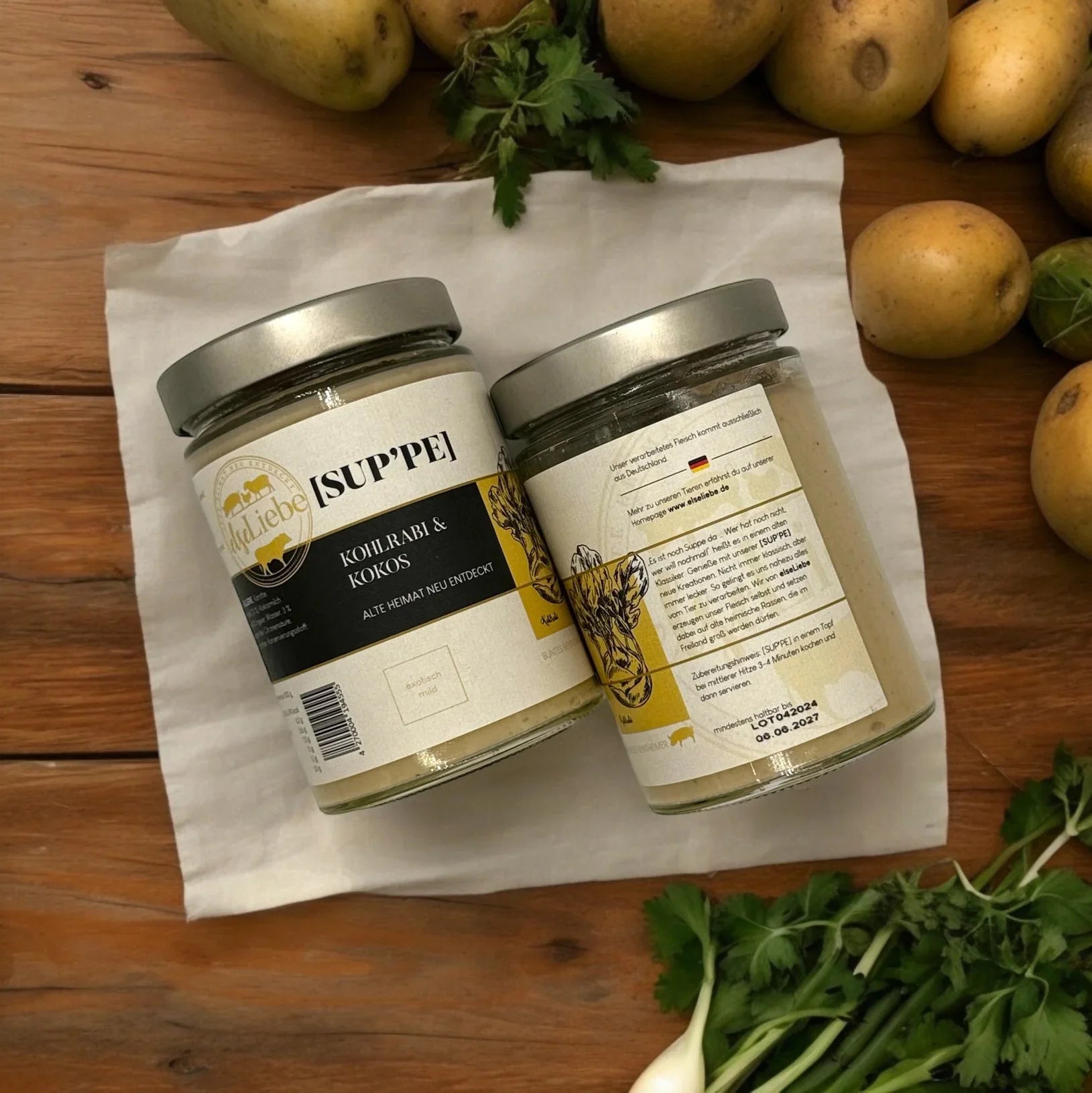 Zwei Gläser Kohlrabi-Kokos-Suppe auf Holz, umgeben von Kartoffeln, Kräutern und Küchenhelfern.