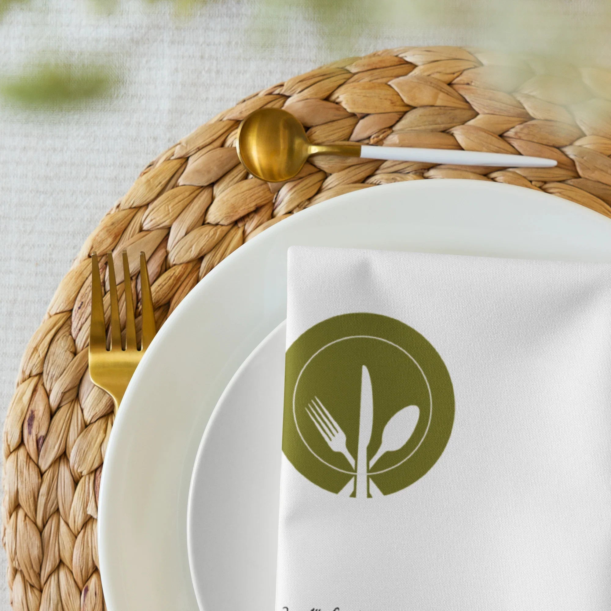 Elegante weiße Serviette mit Besteckmotiv auf geflochtenem Platzset, goldenem Besteck und weißem Teller, stilvolle Küchendeko von Kochschein